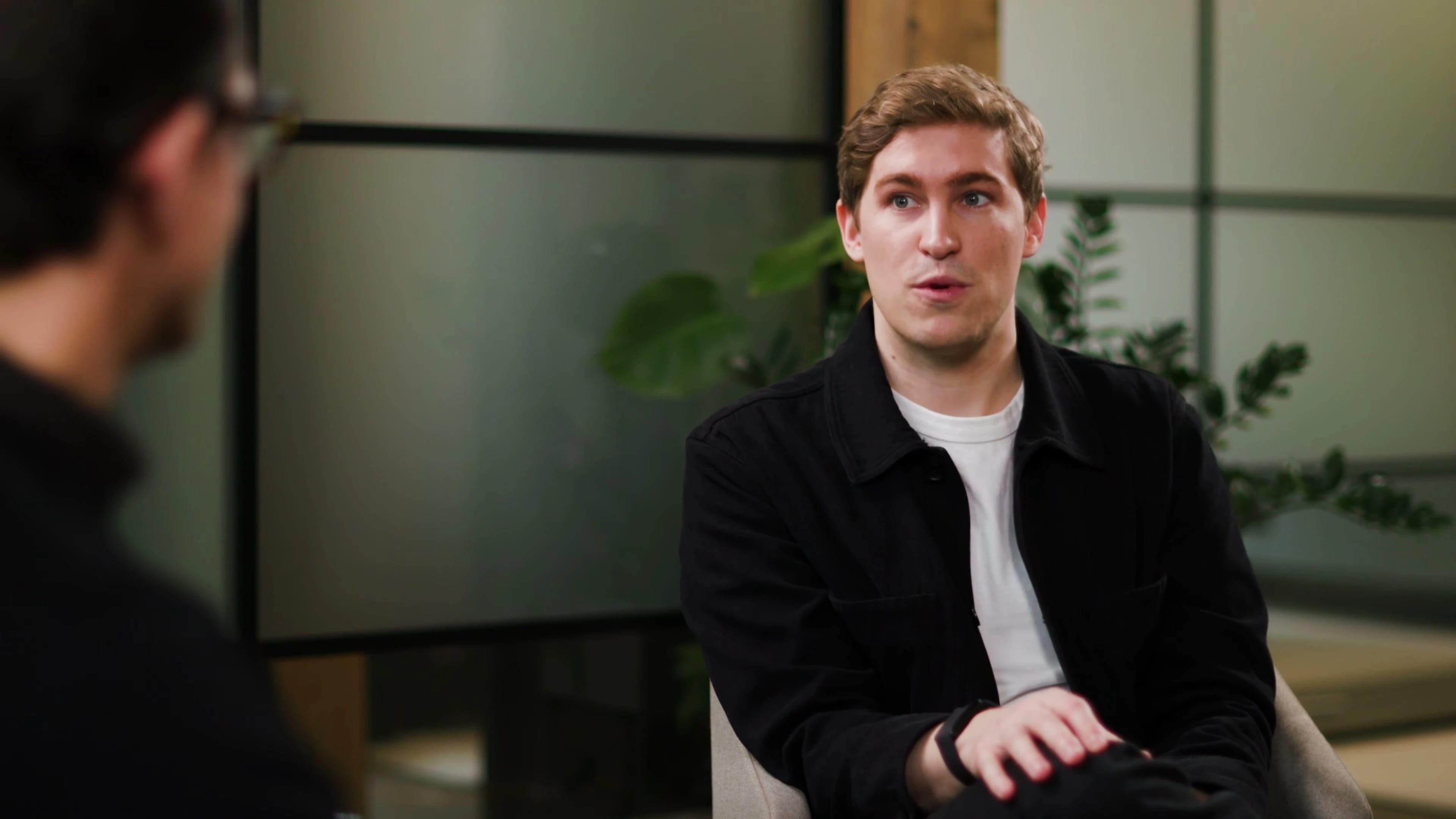 Person speaking in a modern office setting with plants and frosted glass.