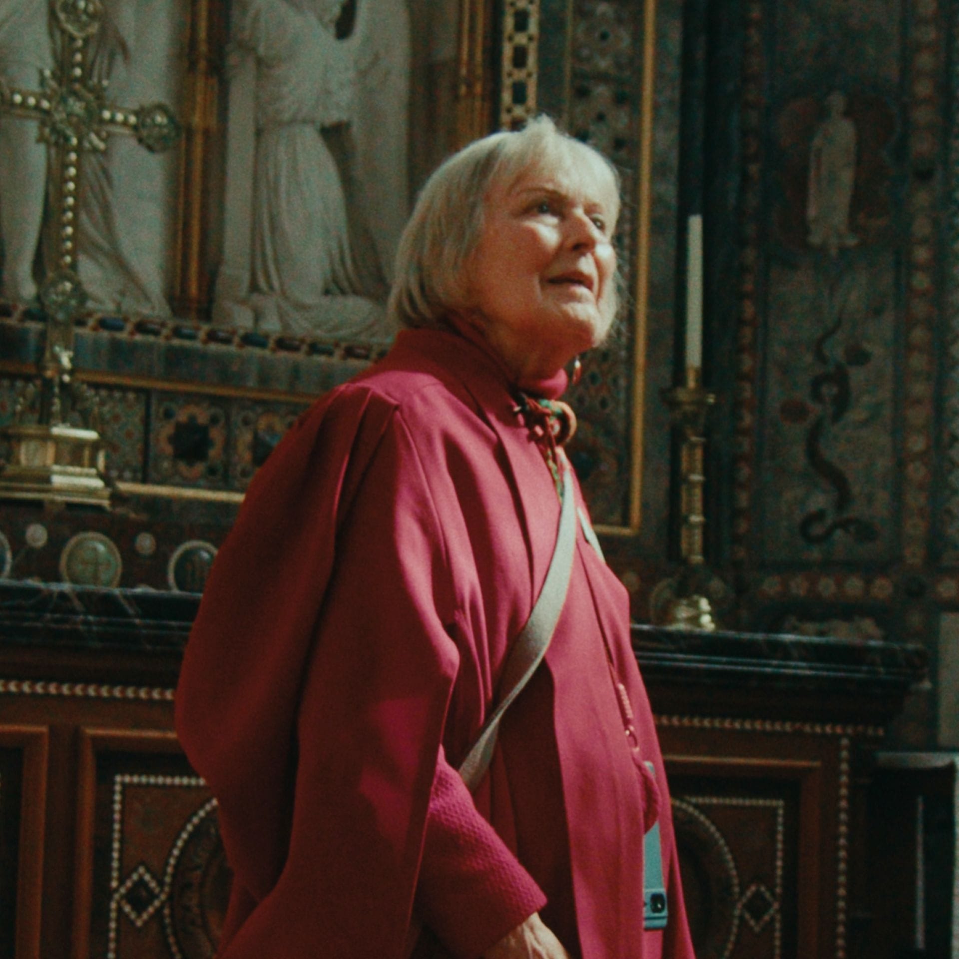 An elderly person in a red robe looks up inside a church, with ornate decorations behind them.