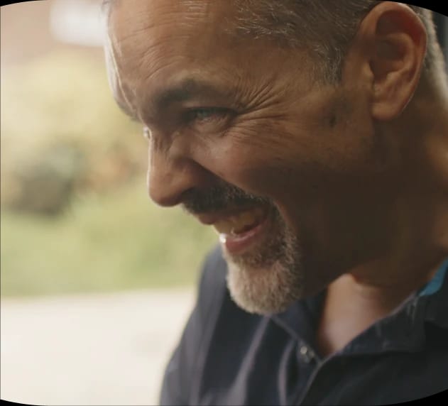 A man smiling broadly, looking to the side, in a natural outdoor setting.
