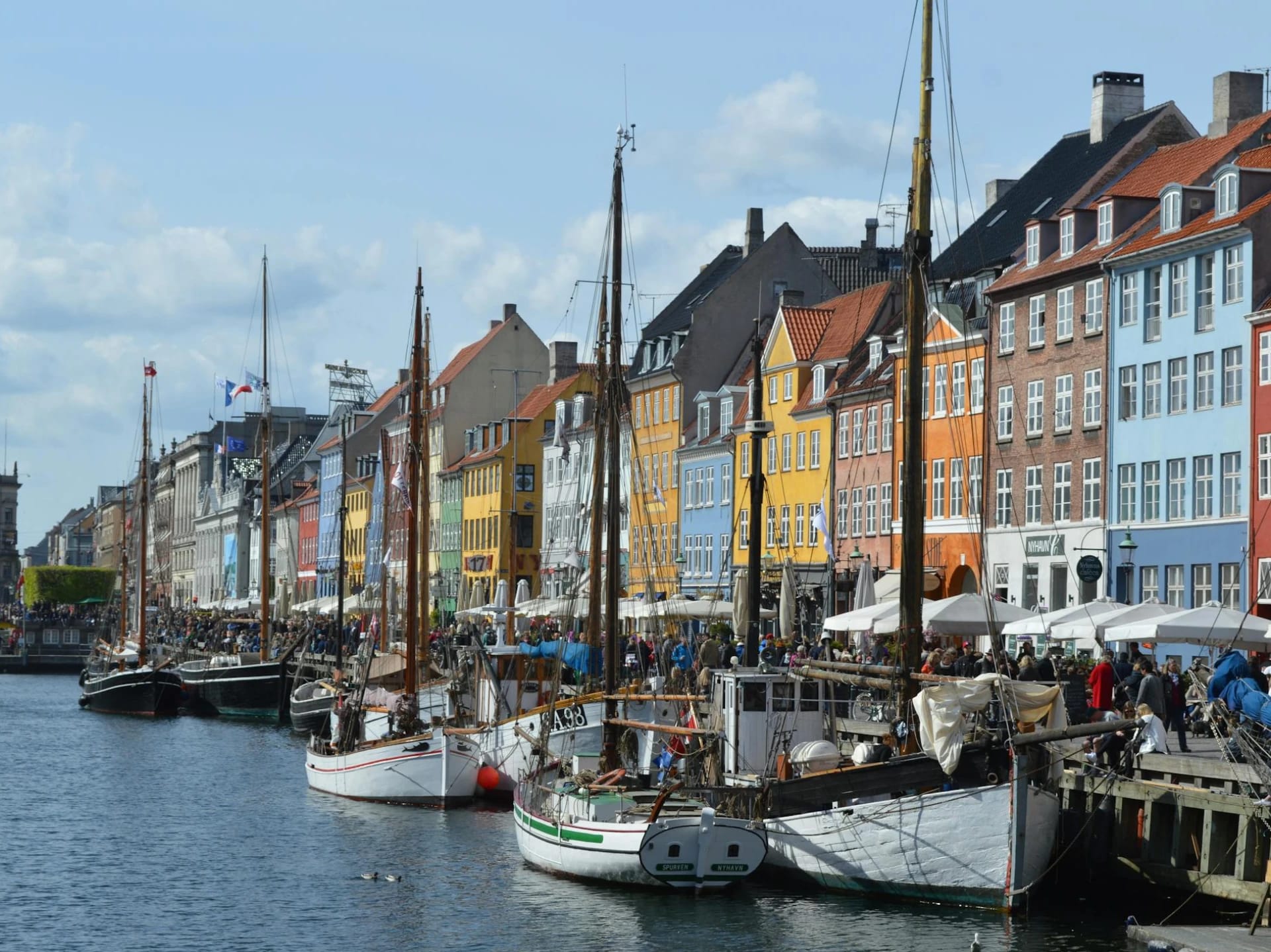 Colorful buildings along a waterfront with sailboats docked in the foreground and people walking on the promenade.