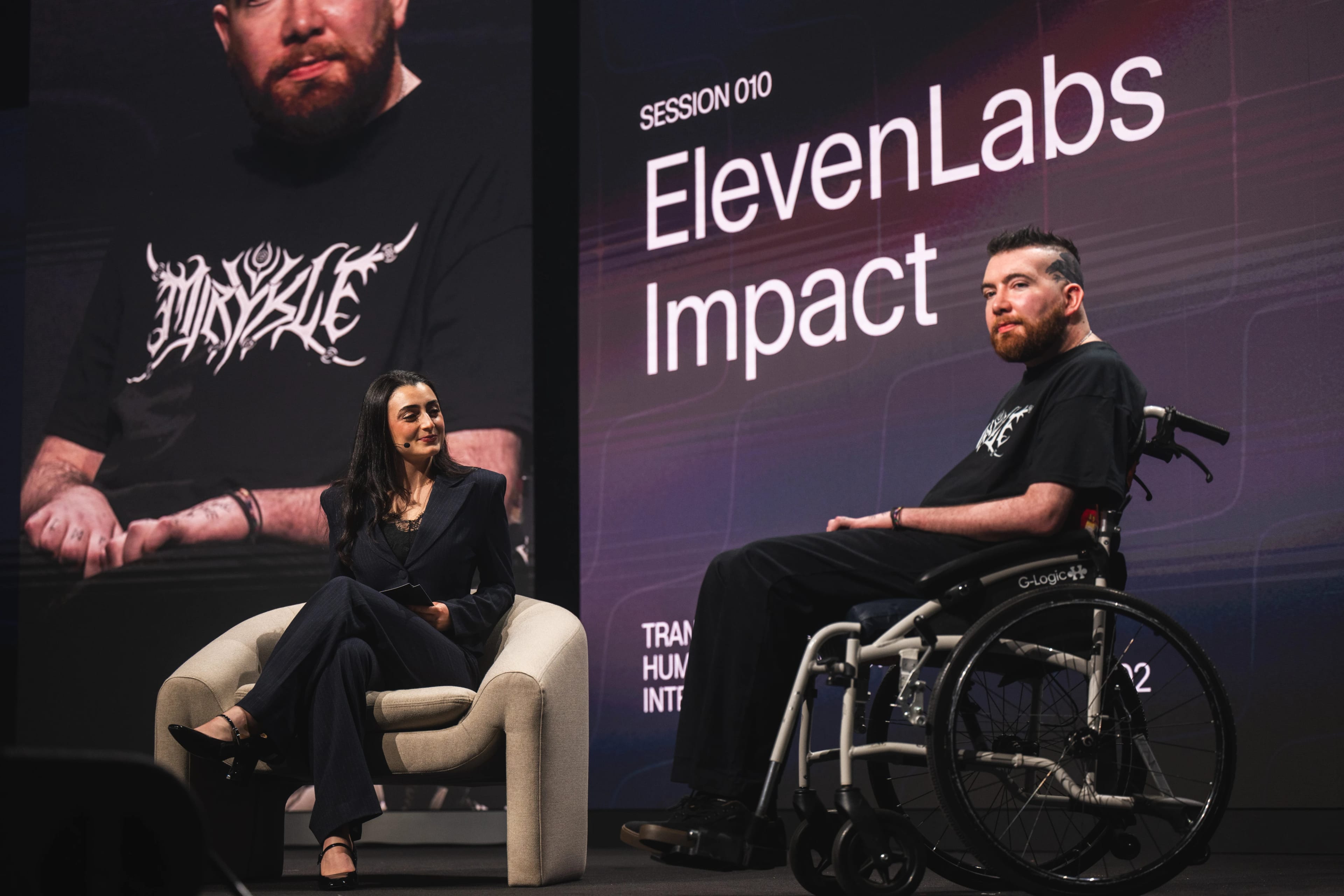 Man in a wheelchair sits on a stage across from a woman sitting in a chair, behind them is a large screen that reads "ElevenLabs Impact"