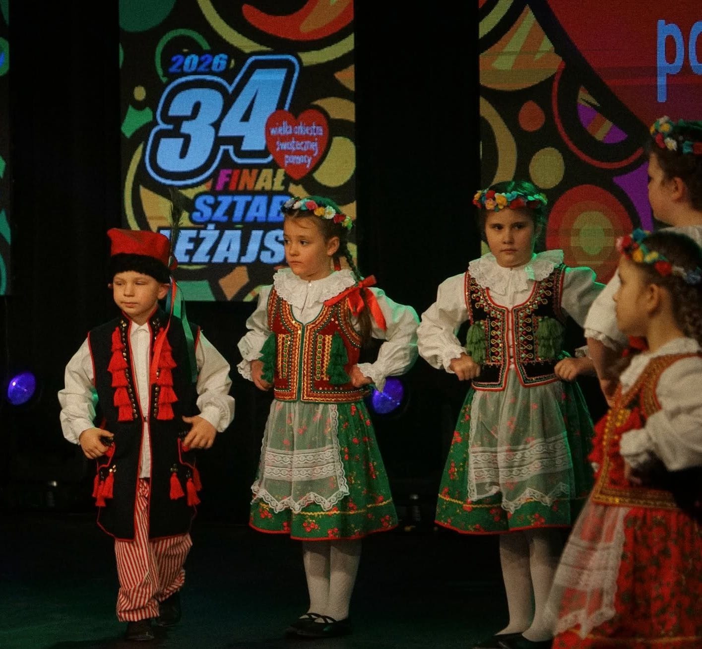 Children in traditional Polish folk costumes performing a dance on stage.