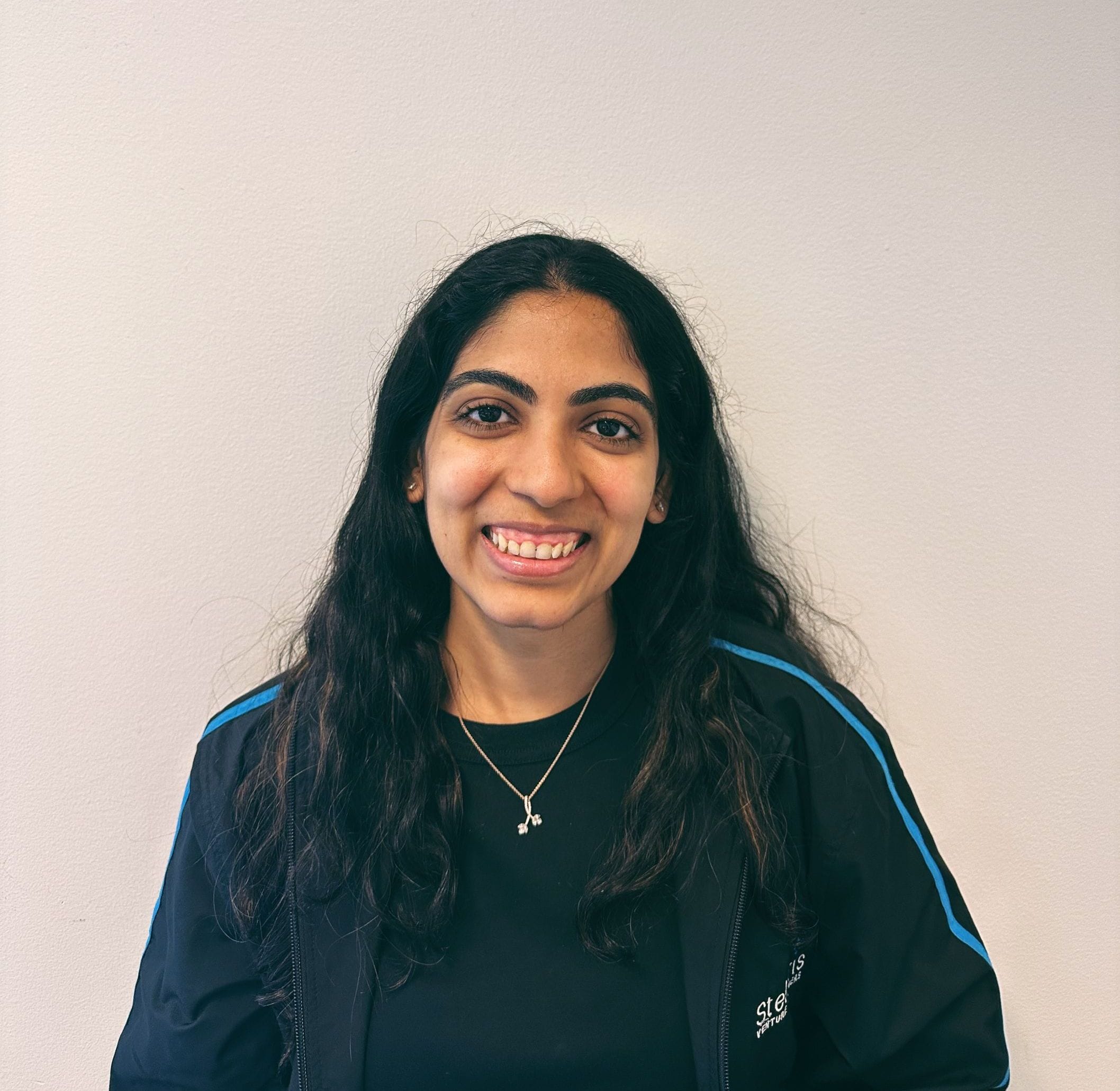 A person smiling against a white wall, wearing a black jacket with blue trim and a necklace.