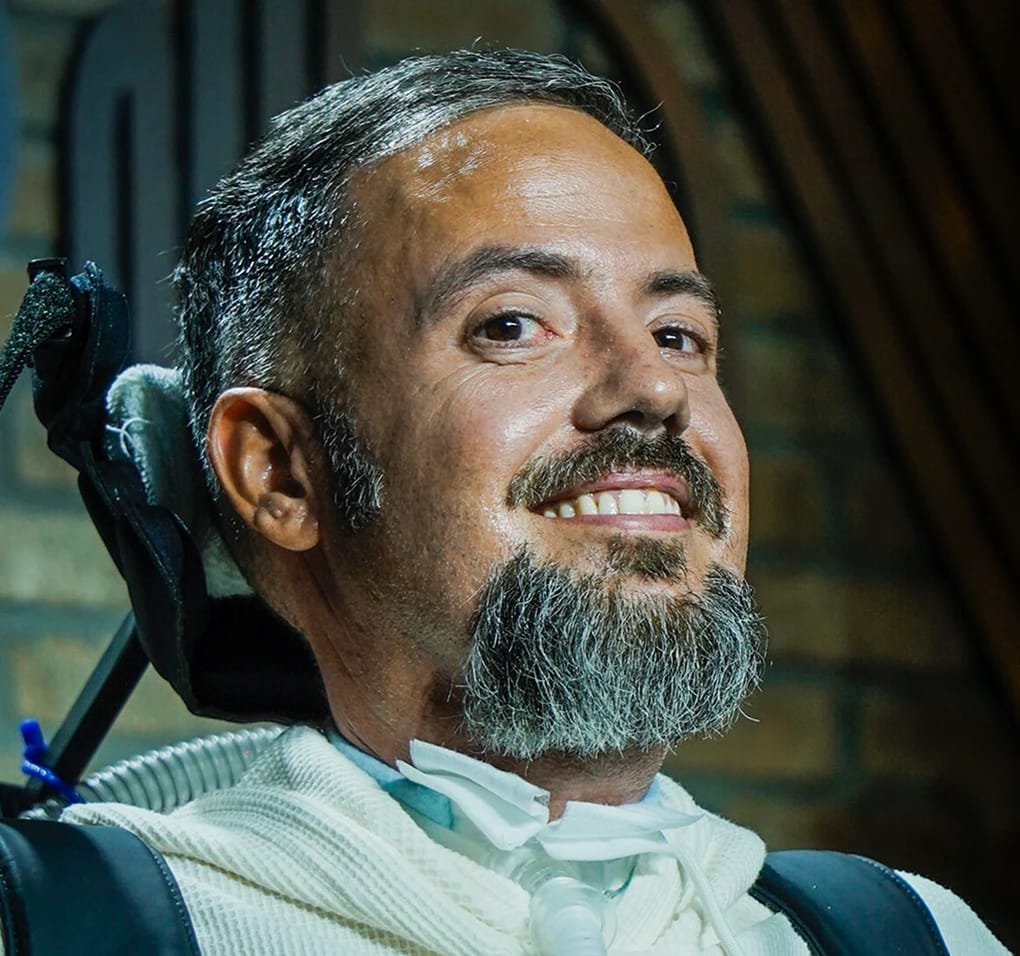 Smiling individual in a wheelchair with a ventilator support device.