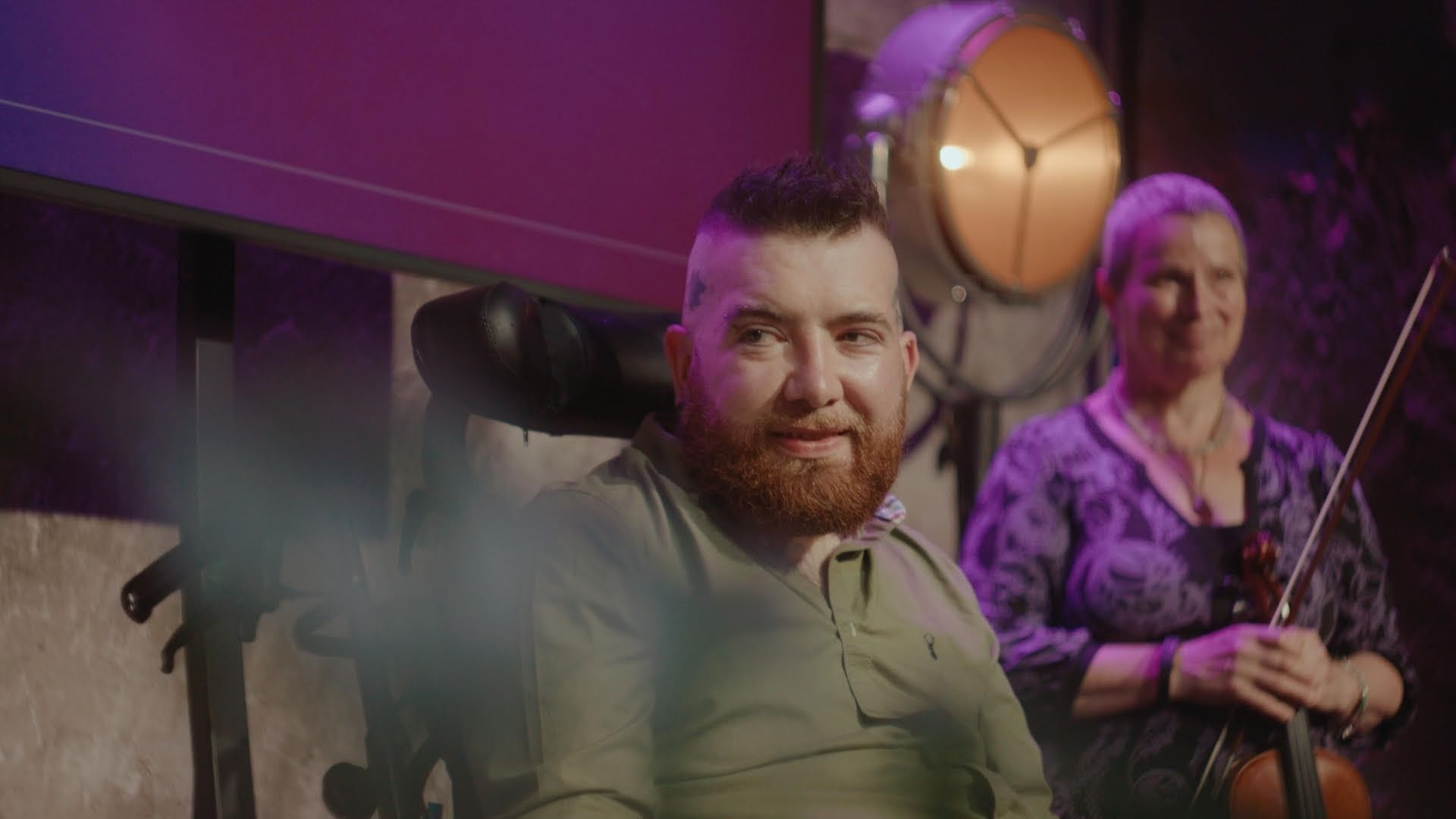 Man with a beanie in a wheelchair smiling next to a woman holding a violin.
