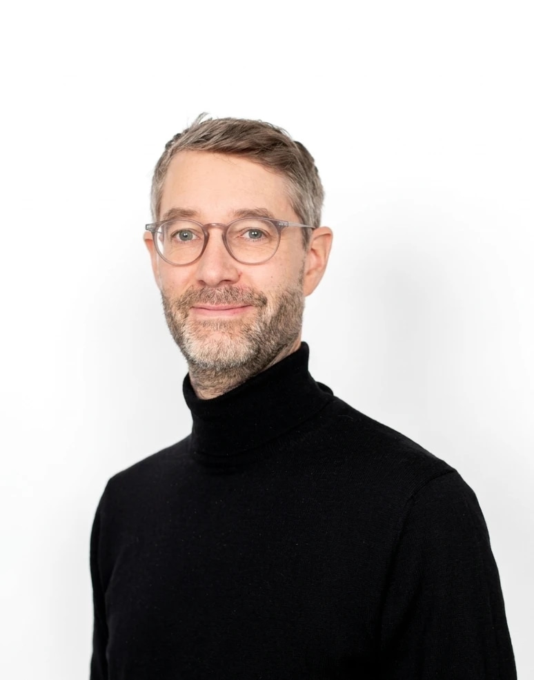 A person with glasses and a black turtleneck, smiling against a white background.