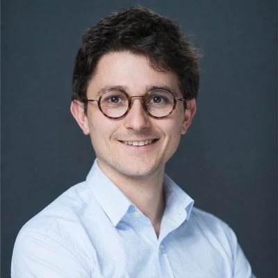 Smiling person with glasses and a light blue shirt against a dark background.