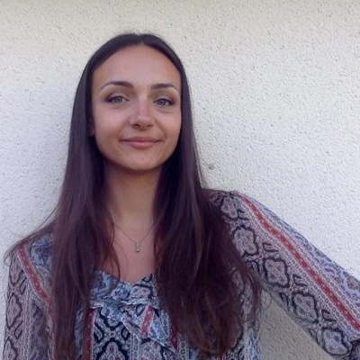 Woman with long hair smiling in front of a plain wall, wearing a patterned blouse.