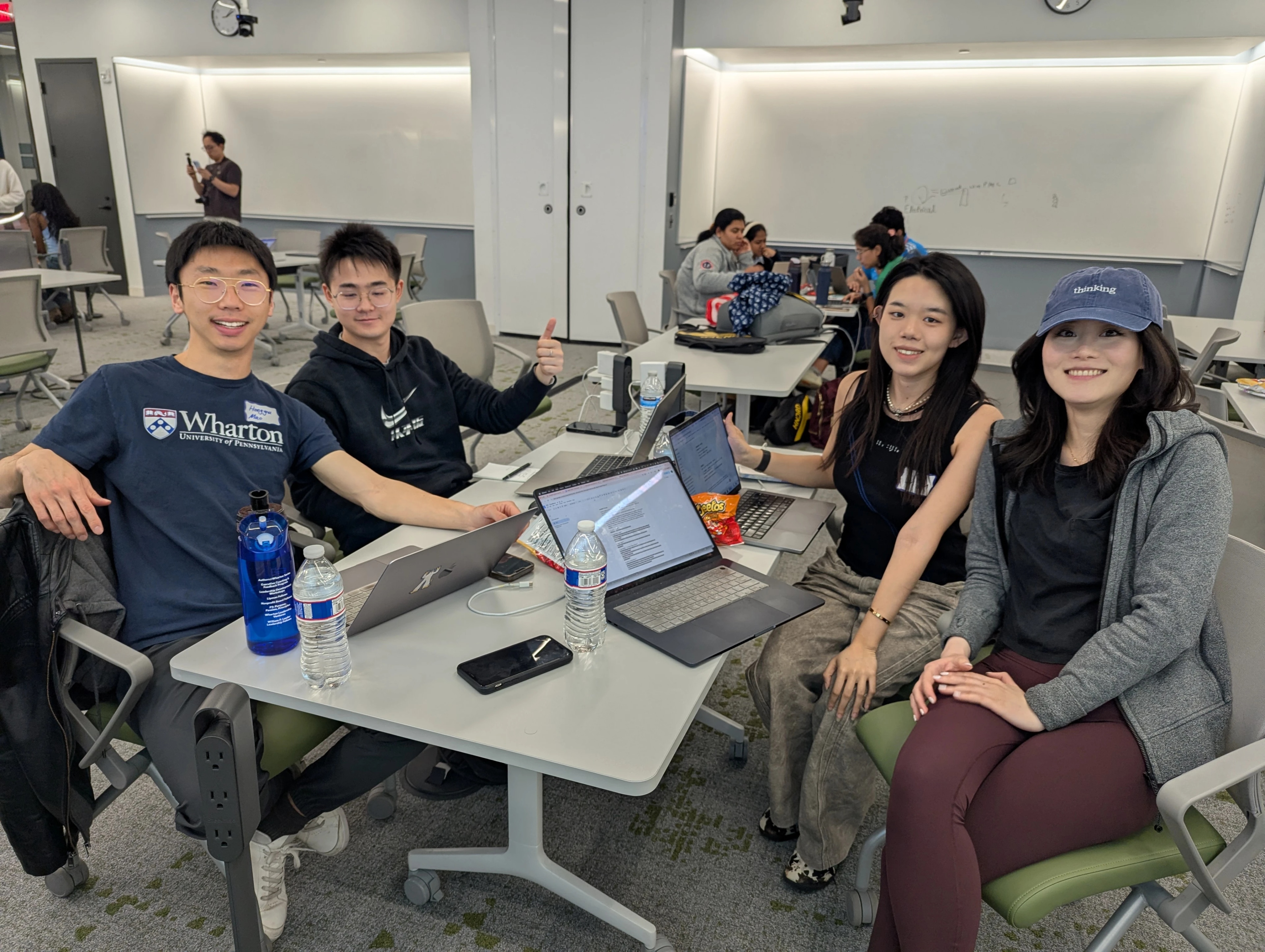 Four people seated at a table in a classroom setting, working on laptops and smiling