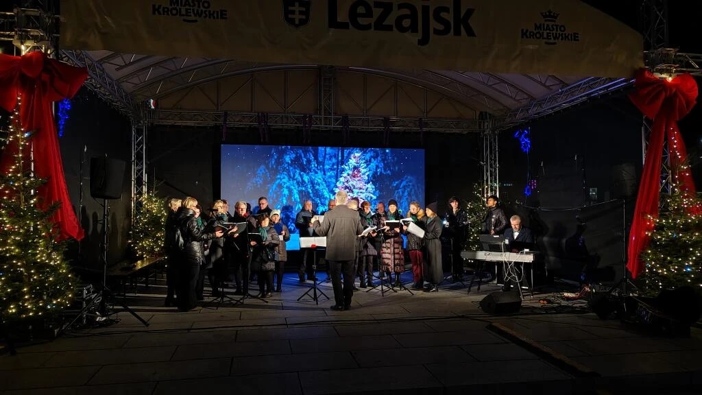A choir performs on an outdoor stage decorated with Christmas lights and trees.