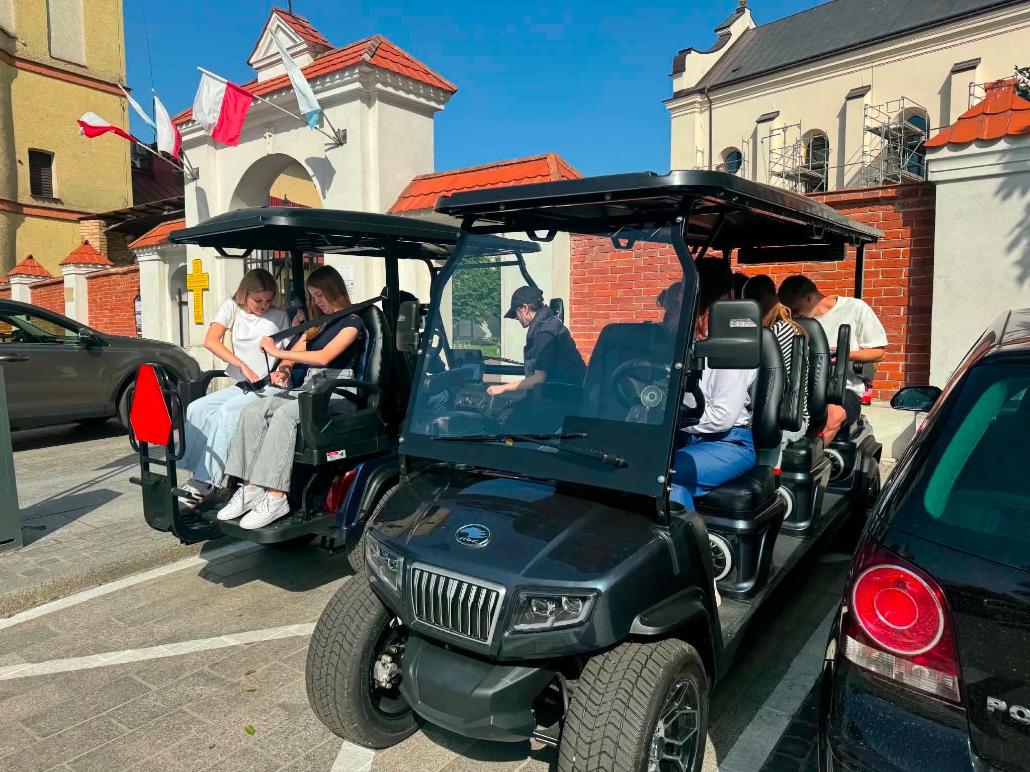People sitting in a golf cart in a sunny parking area near a brick building with flags.