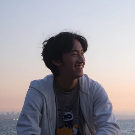 A person smiling by the ocean at sunset, wearing a raccoon graphic t-shirt and a light jacket.iel