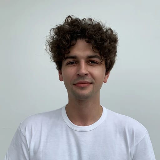 A young man with curly hair wearing a white t-shirt, smiling softly against a plain background.