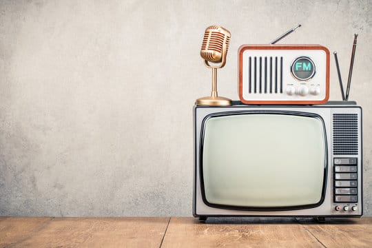 Vintage radio and microphone on top of an old television set against a plain wall.