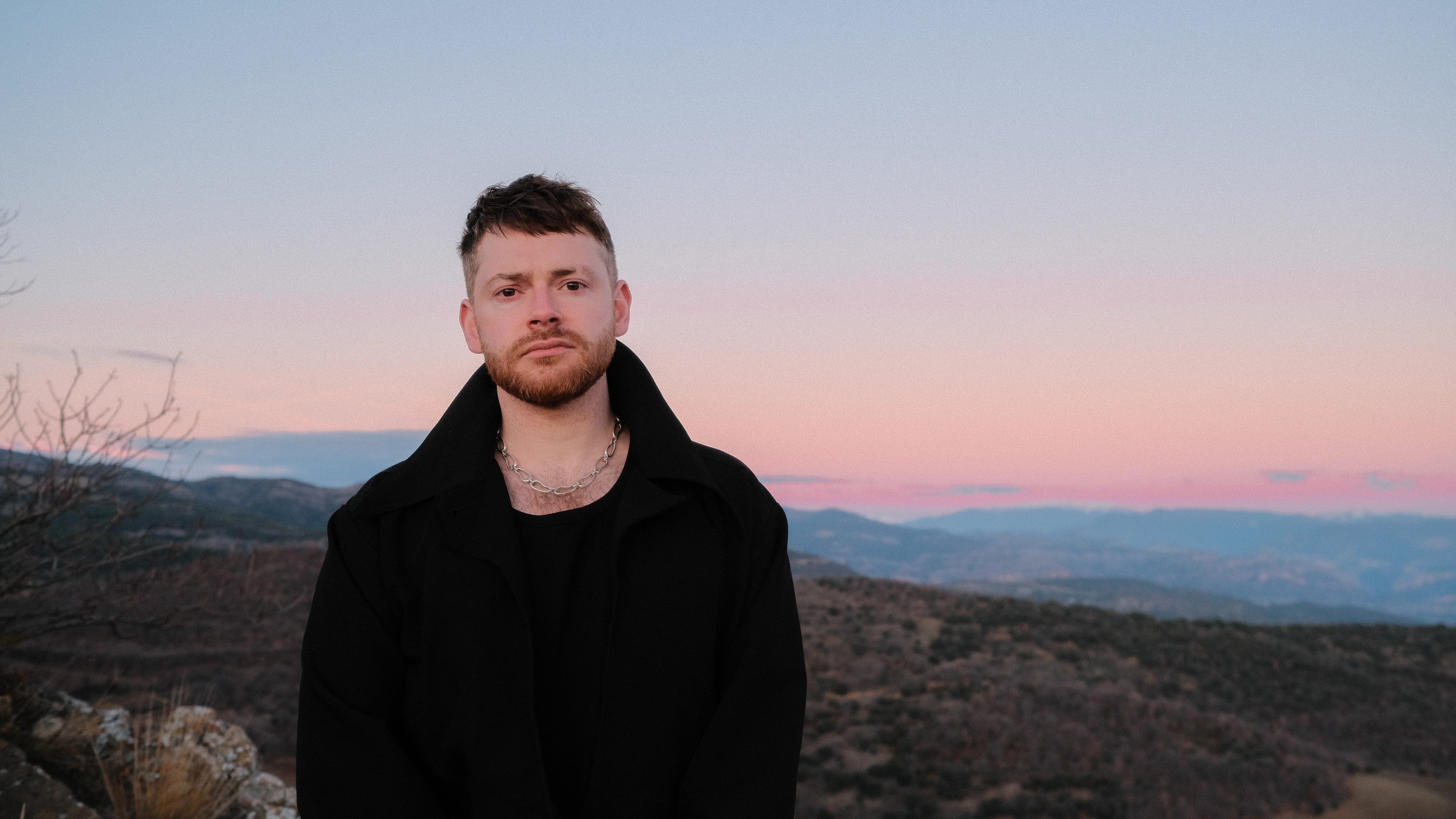 A man with short hair and a beard standing outdoors during sunset, wearing a black jacket and a silver chain necklace, with a landscape of mountains and a pink and blue sky in the background.